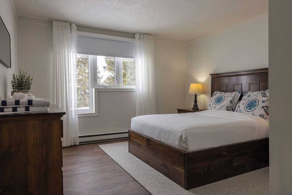 A bedroom with a wooden bed, a nightstand with a lamp, a window with curtains, and a plant on a dresser.