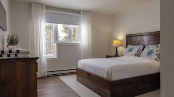 A bedroom with a wooden bed, a nightstand with a lamp, a window with curtains, and a plant on a dresser.