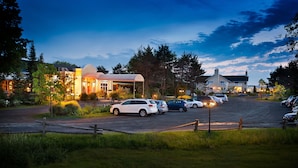 A parking lot with several cars and a building with a covered area. - Auberge West Brome (Lac-Brome)