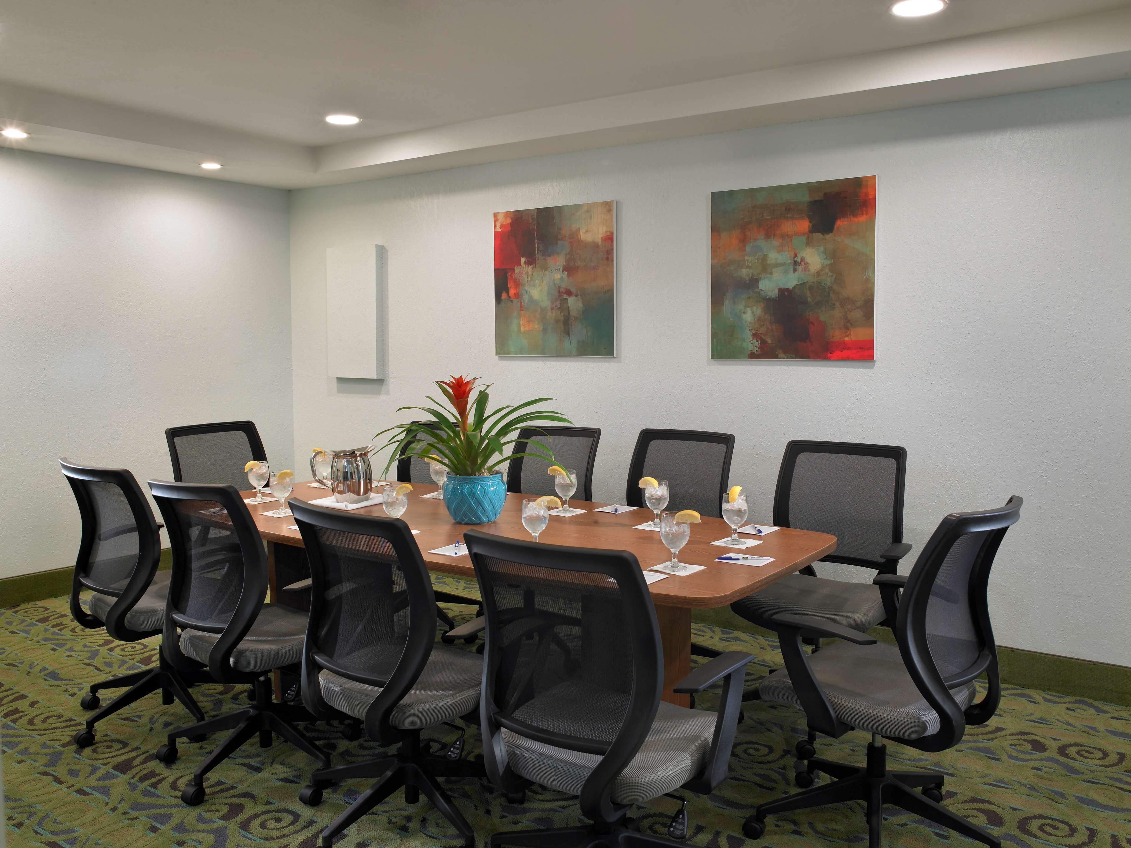 A conference room with a wooden table, black mesh chairs, a blue vase with a plant, and two abstract paintings on the wall.