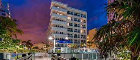 A multi-story hotel with balconies, a pool area with lounge chairs, and a wooden bridge leading to the pool.