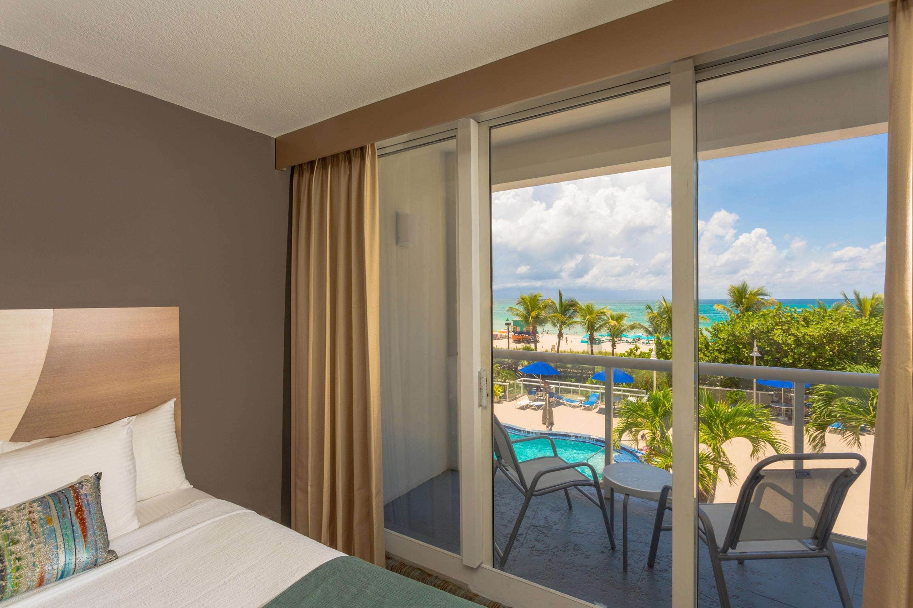 A hotel room with a bed, a view of a pool and beach, and a balcony with chairs.