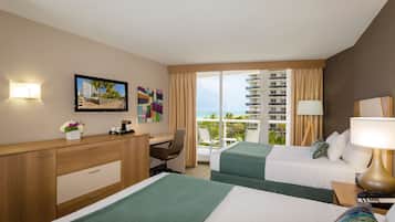 Quarto de hotel com duas camas, uma mesa, uma cadeira, um abajur e vista para o mar.