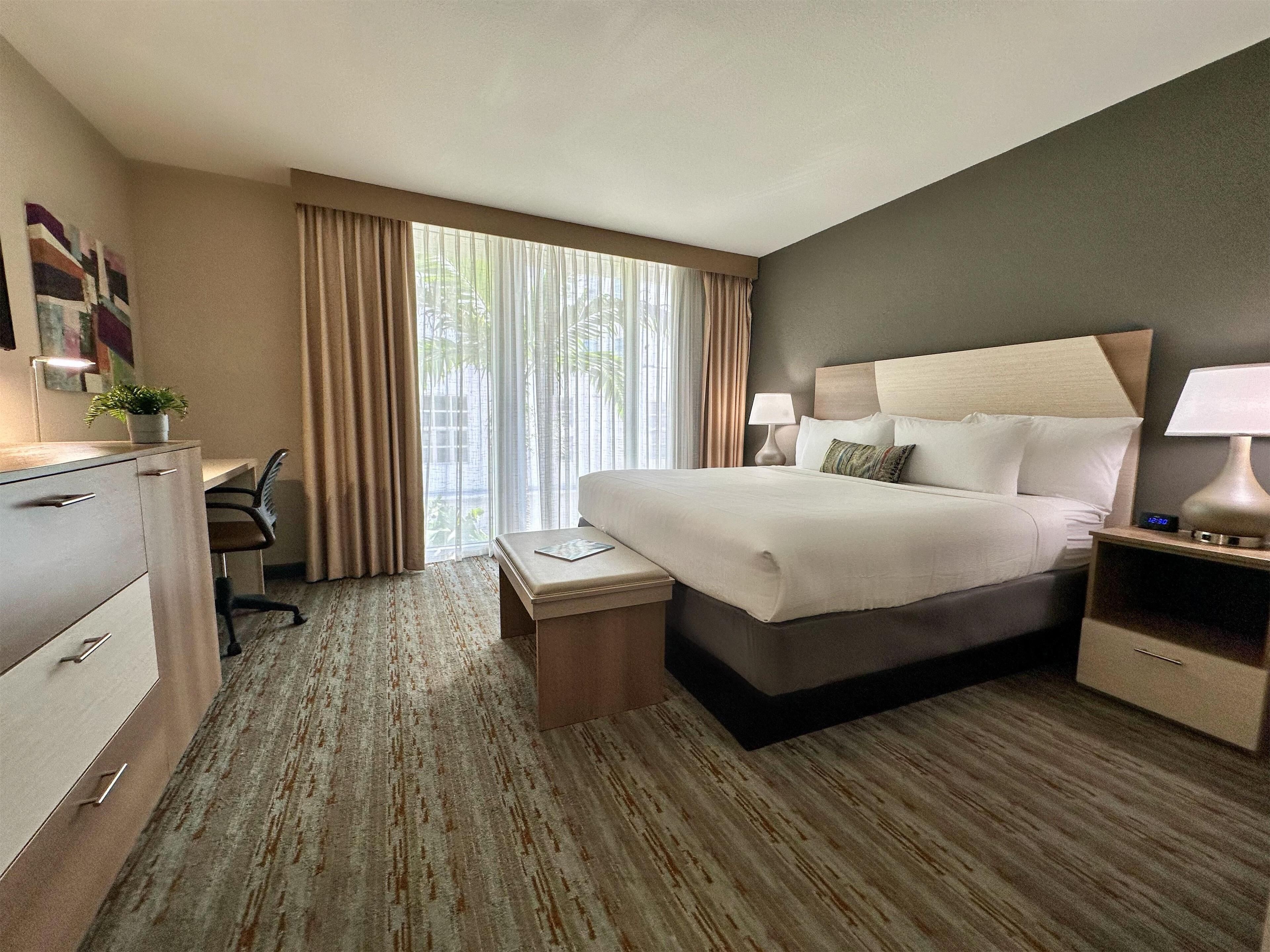 A hotel room with a large bed, a desk with a chair, a small table, and a nightstand with a lamp.