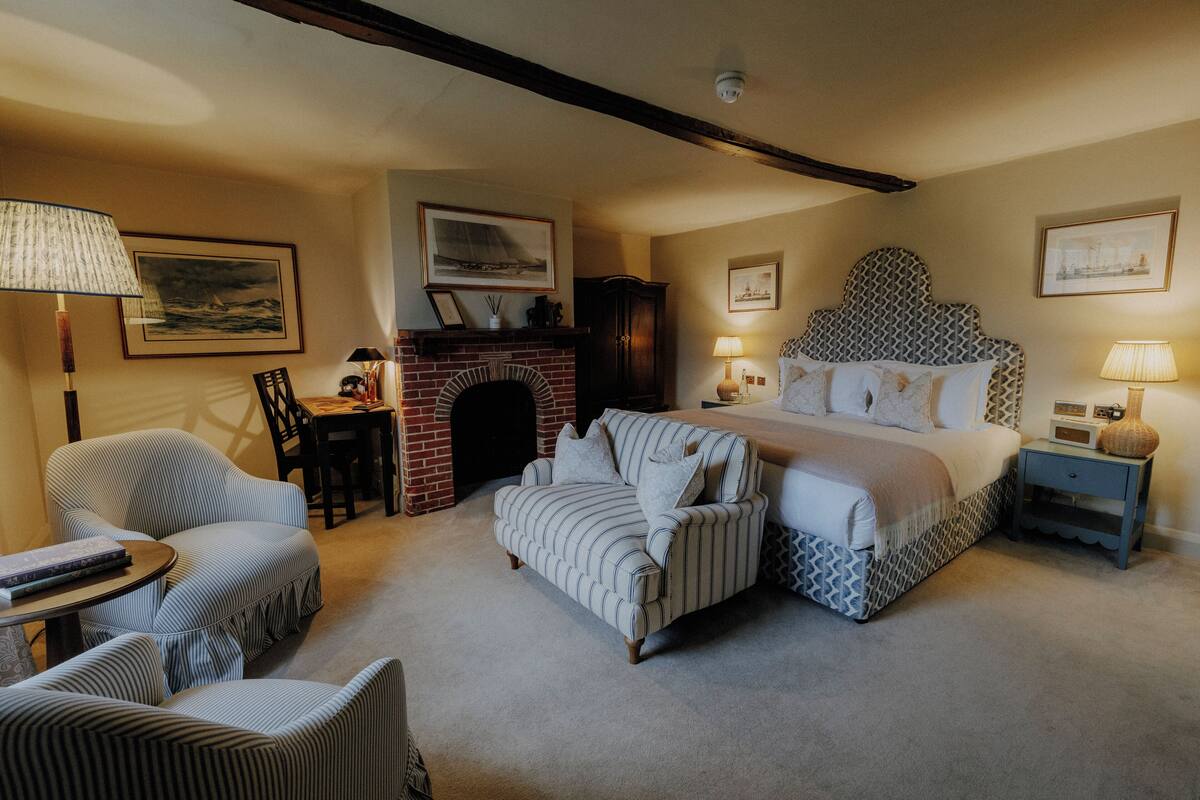 A bedroom with a large bed, two armchairs, a small table, a fireplace, and a nightstand with lamps.