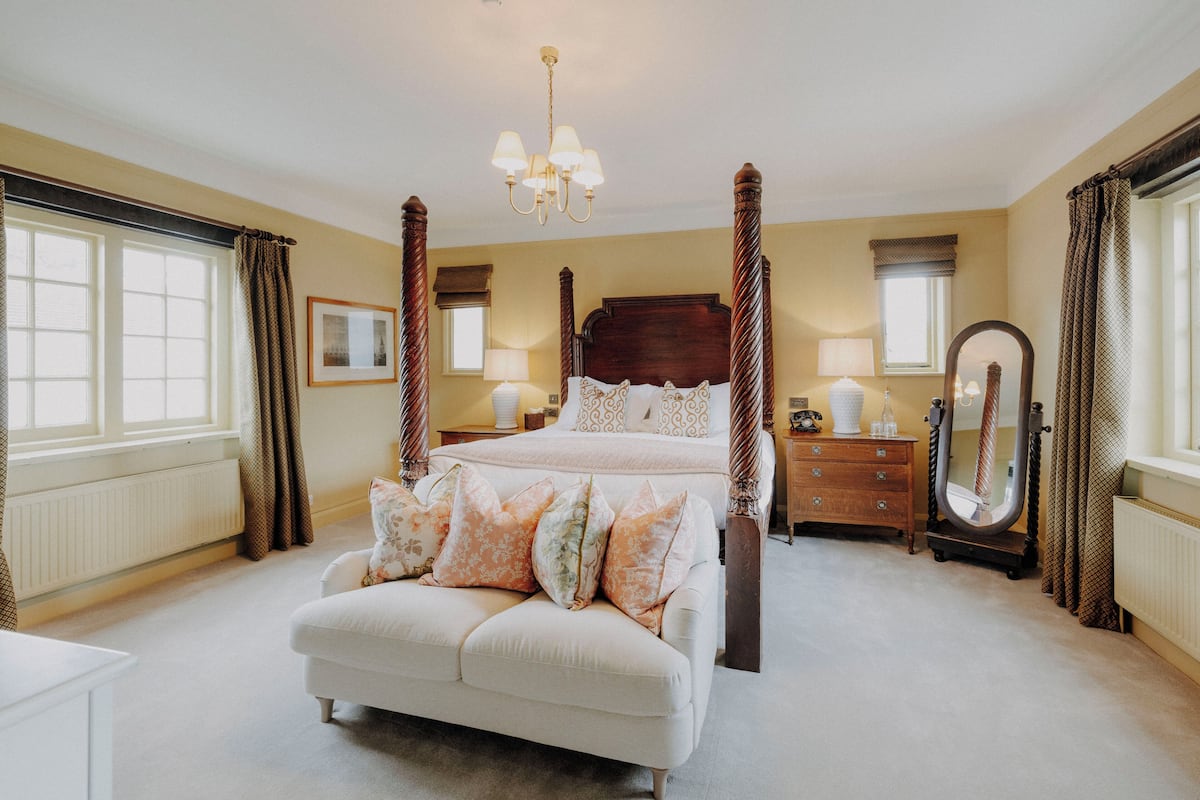 A bedroom with a four-poster bed, a sitting area, a dresser, a mirror, and a chandelier.