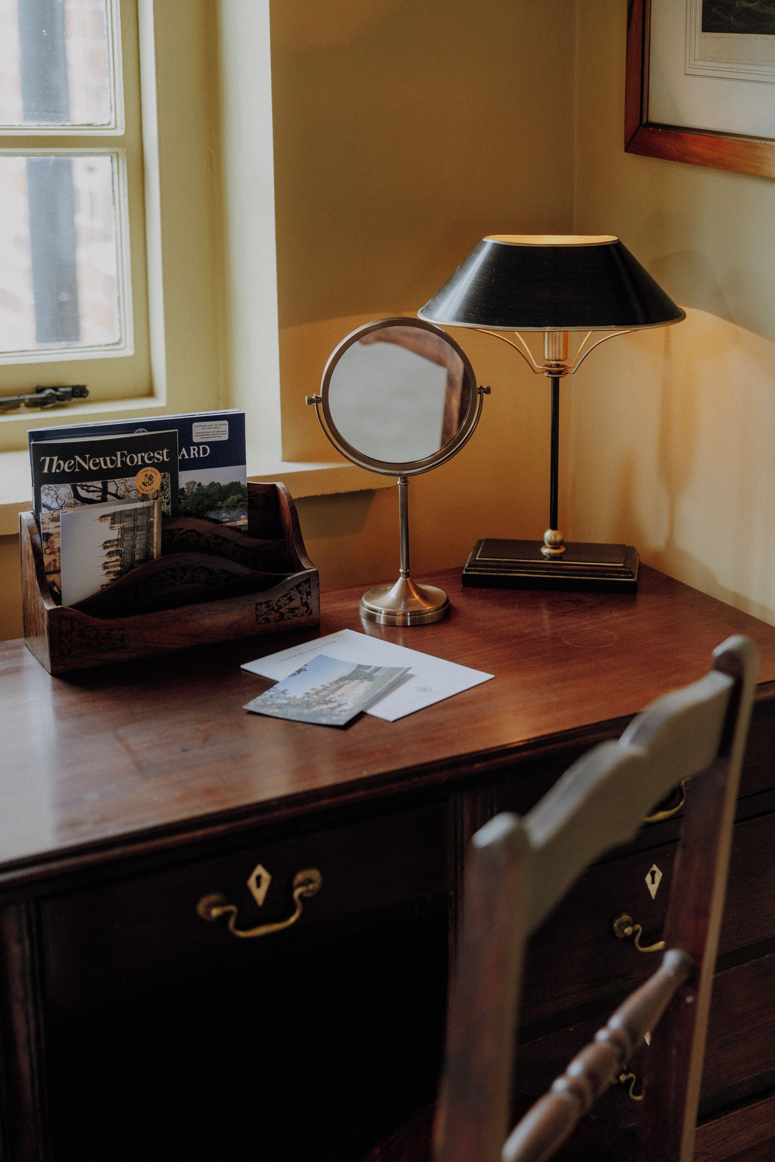 A wooden desk with a lamp, a mirror, a book, and a chair.