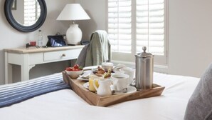 A neatly made bed with a tray of breakfast items, a bedside table with a lamp, a mirror, and a window with blinds. - Master Builders House Hotel (Brockenhurst)