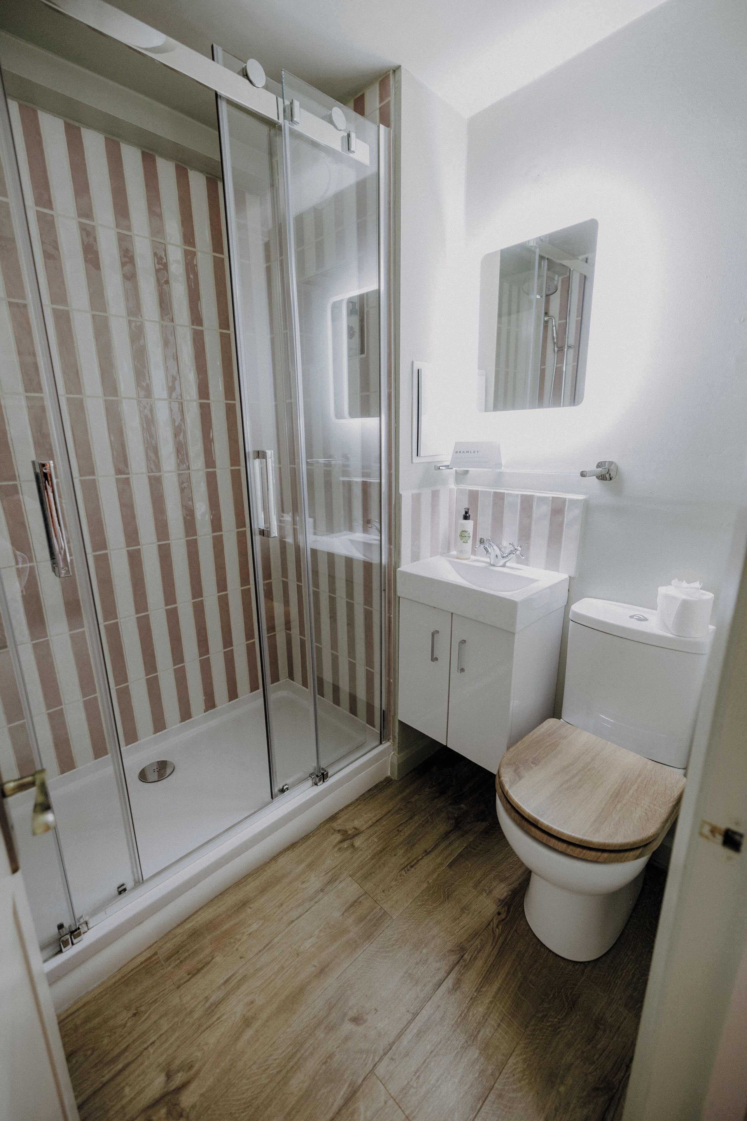 Un baño con cabina de ducha de vidrio, inodoro de madera, lavamanos con espejo y azulejos a rayas en las paredes.