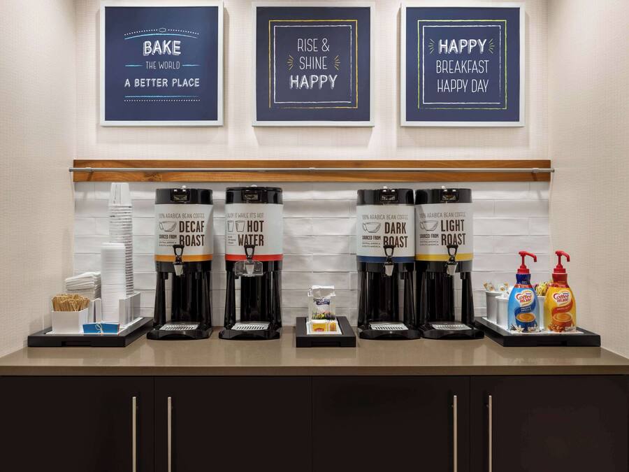 A coffee station with multiple coffee dispensers labeled 'Decaf', 'Hot', 'Dark', and 'Light'. Above the station, there are three framed signs with motivational messages.