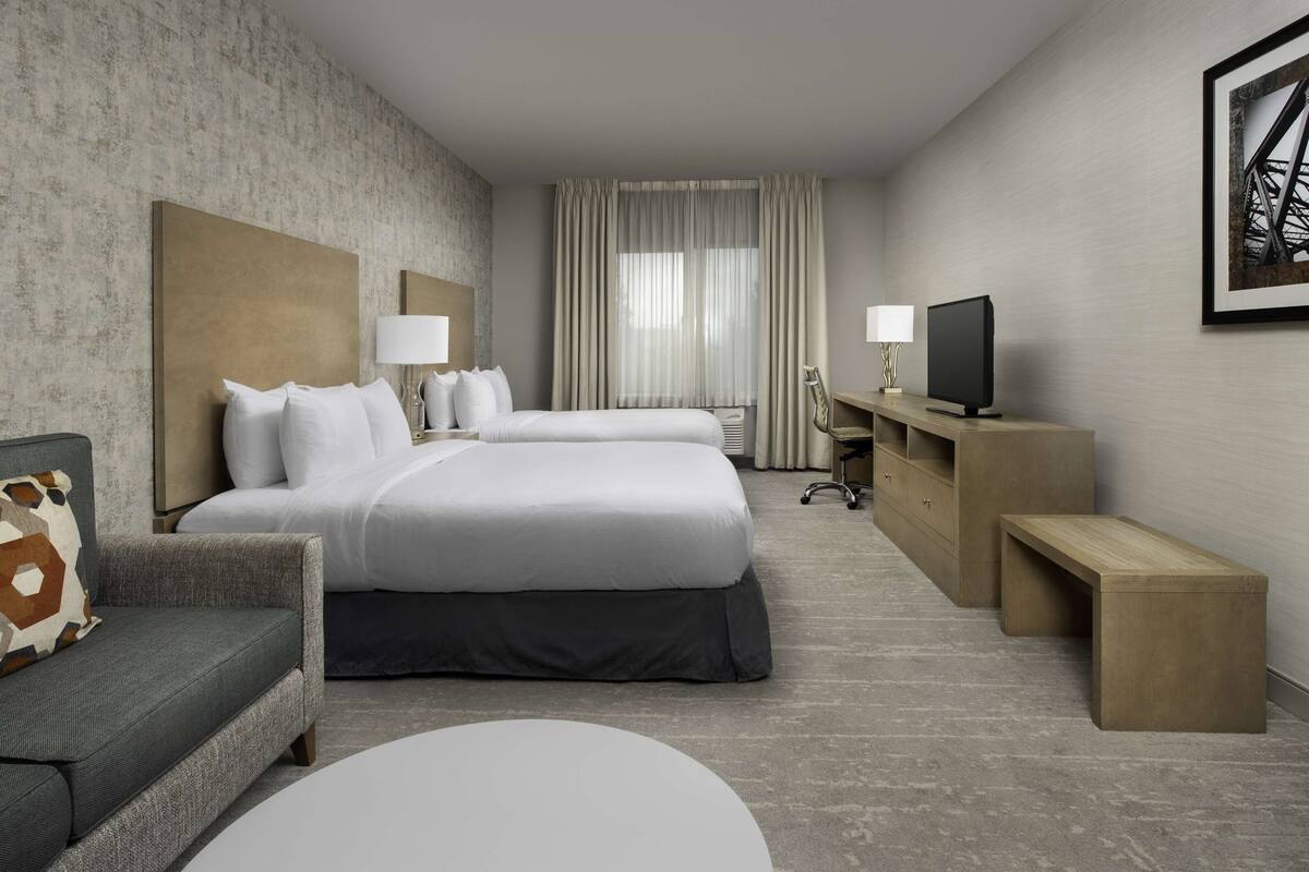 A hotel room with two beds, a sofa, a desk with a TV, and a large window with curtains.