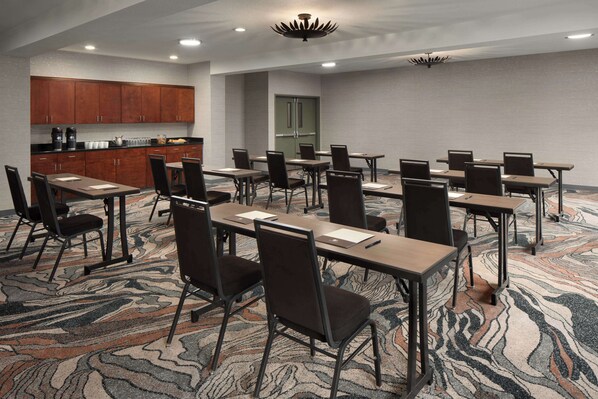 A conference room with multiple tables and chairs, a kitchen area with wooden cabinets, and a patterned carpet.