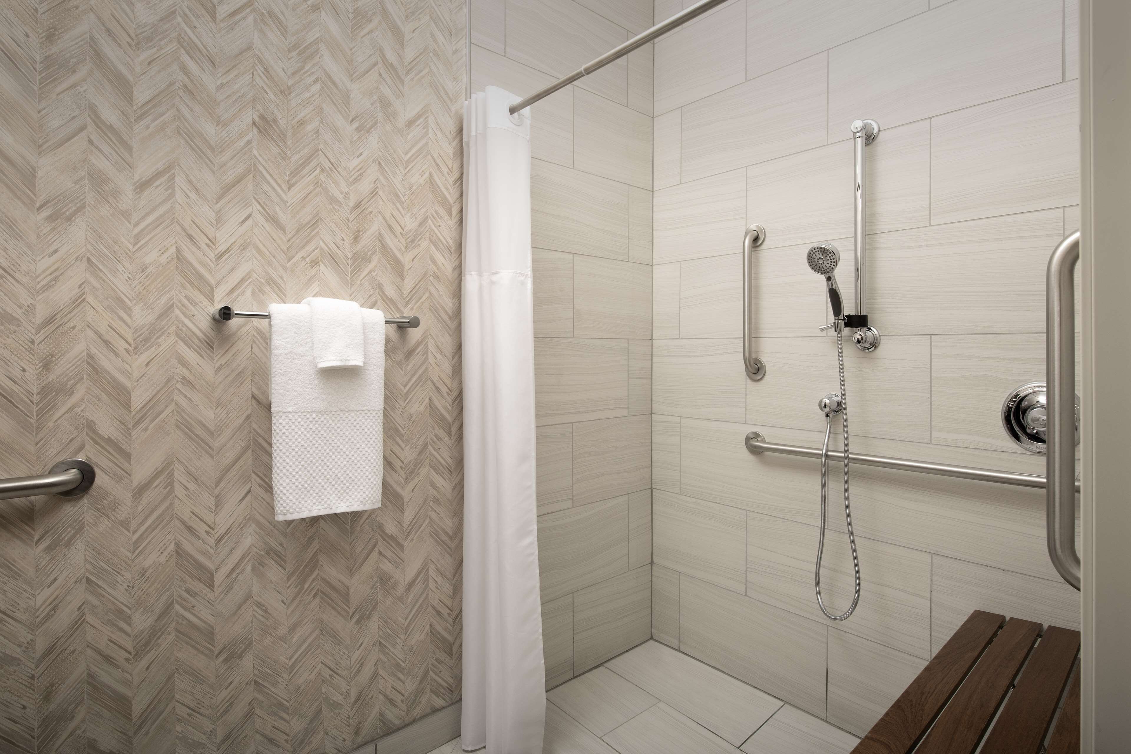 A shower area with a herringbone-patterned wall, a white towel hanging on a metal rack, a showerhead, and a wooden bench.