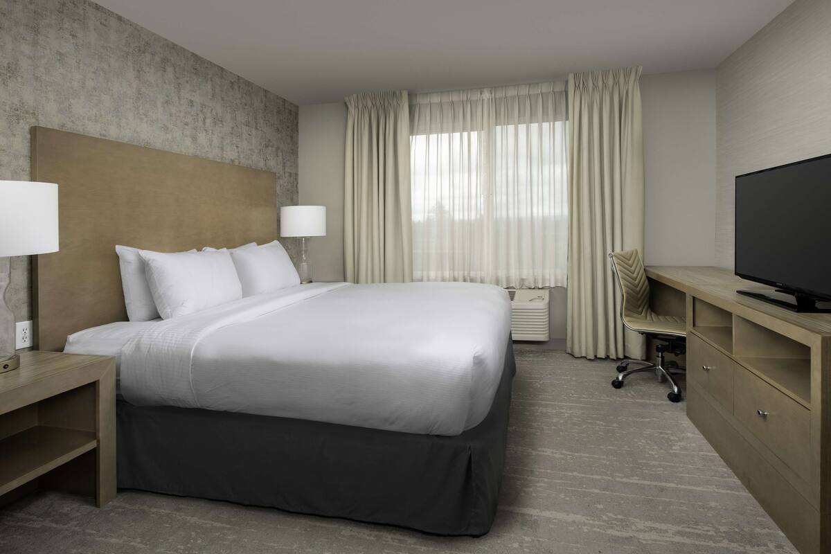 A hotel room with a large bed, bedside tables with lamps, a desk with a TV, and a window with curtains.