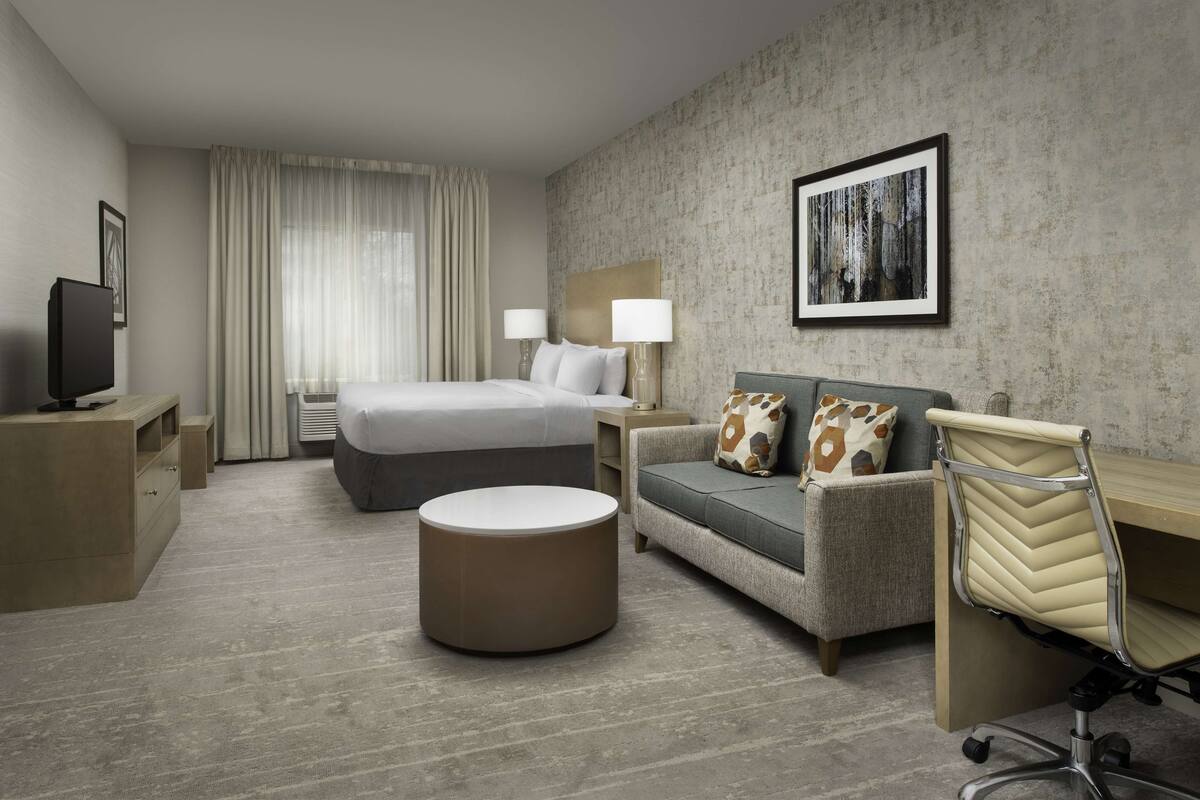 A modern hotel room with a bed, a round ottoman, a sofa, a desk, and a chair.