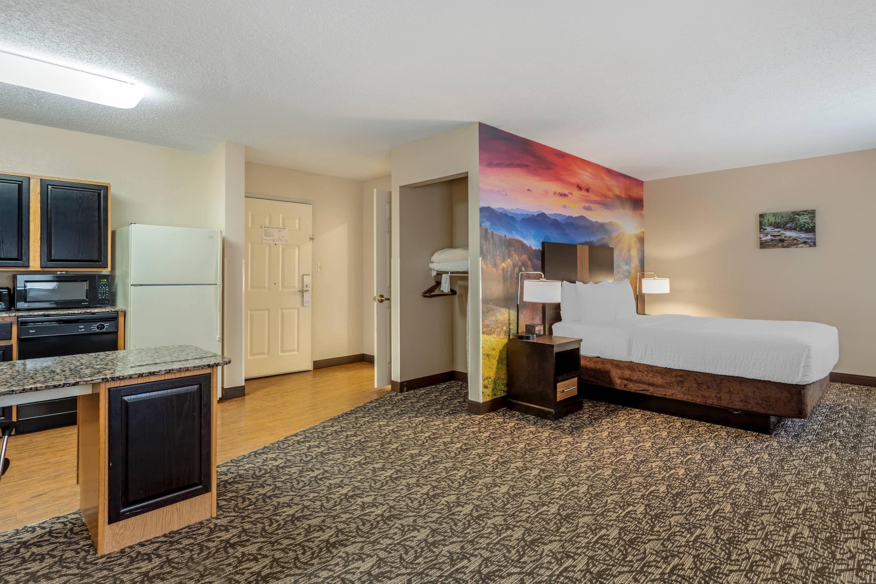 A hotel room with a bed, a desk, a microwave, a refrigerator, and a painting of a sunset.