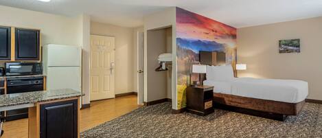 A hotel room with a bed, a desk, a microwave, a refrigerator, and a painting of a sunset.