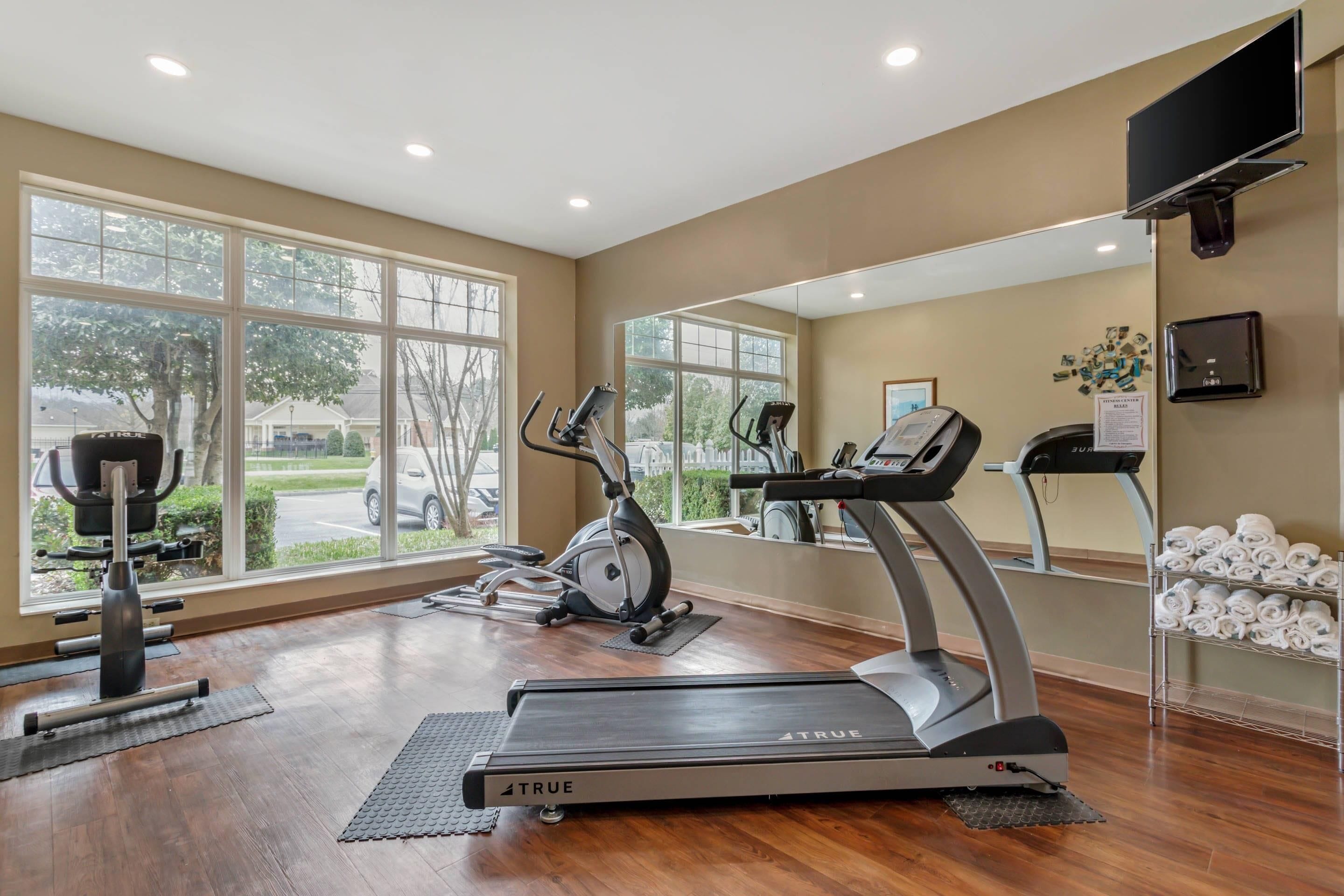 A gym with treadmills, elliptical machines, and weight equipment. Large windows offer views of the outdoors.