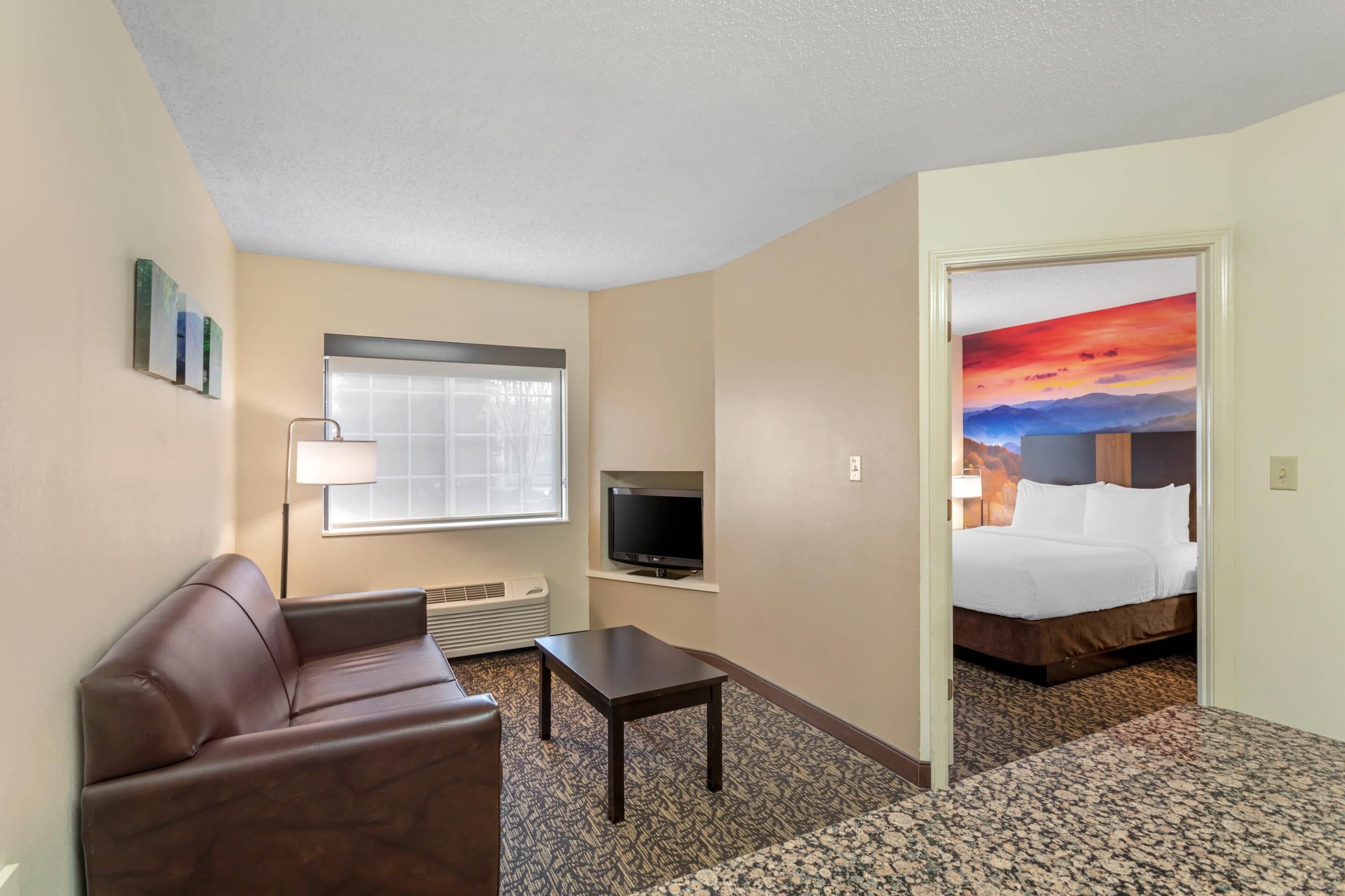 A hotel room with a sofa, a TV, a small table, a window, and a bed with a painting on the wall.