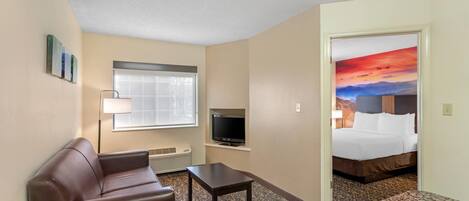 A hotel room with a sofa, a TV, a small table, a window, and a bed with a painting on the wall.