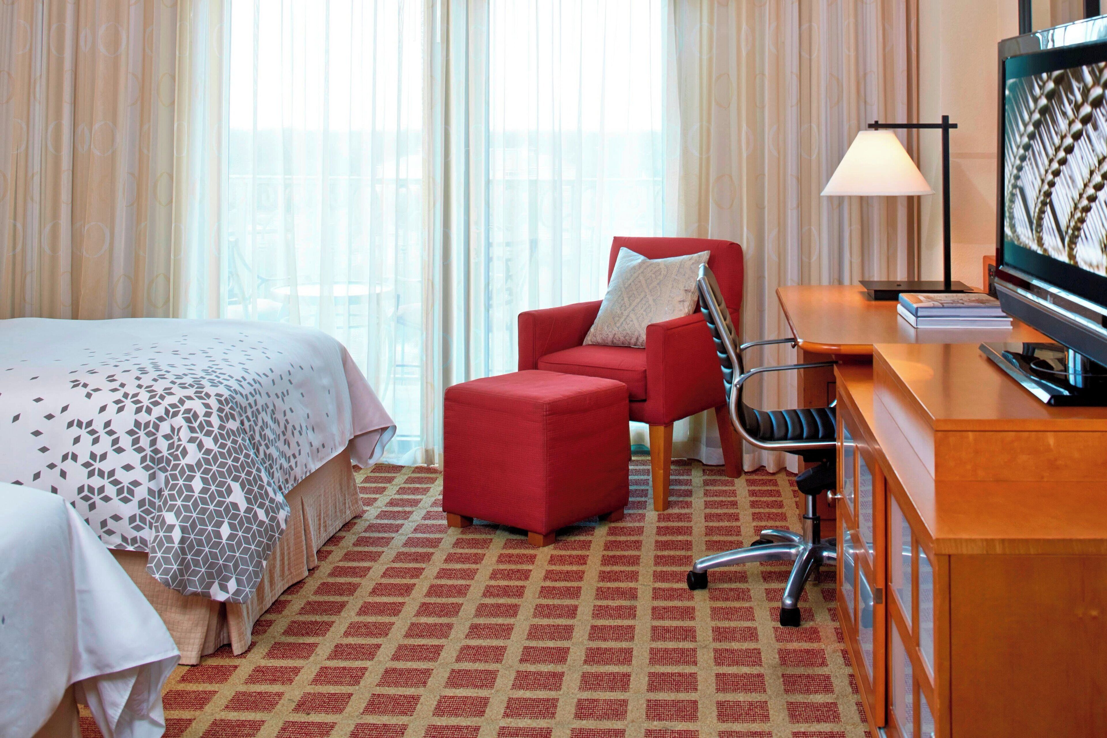 A hotel room with a bed, a red armchair, a desk with a computer, and a lamp.