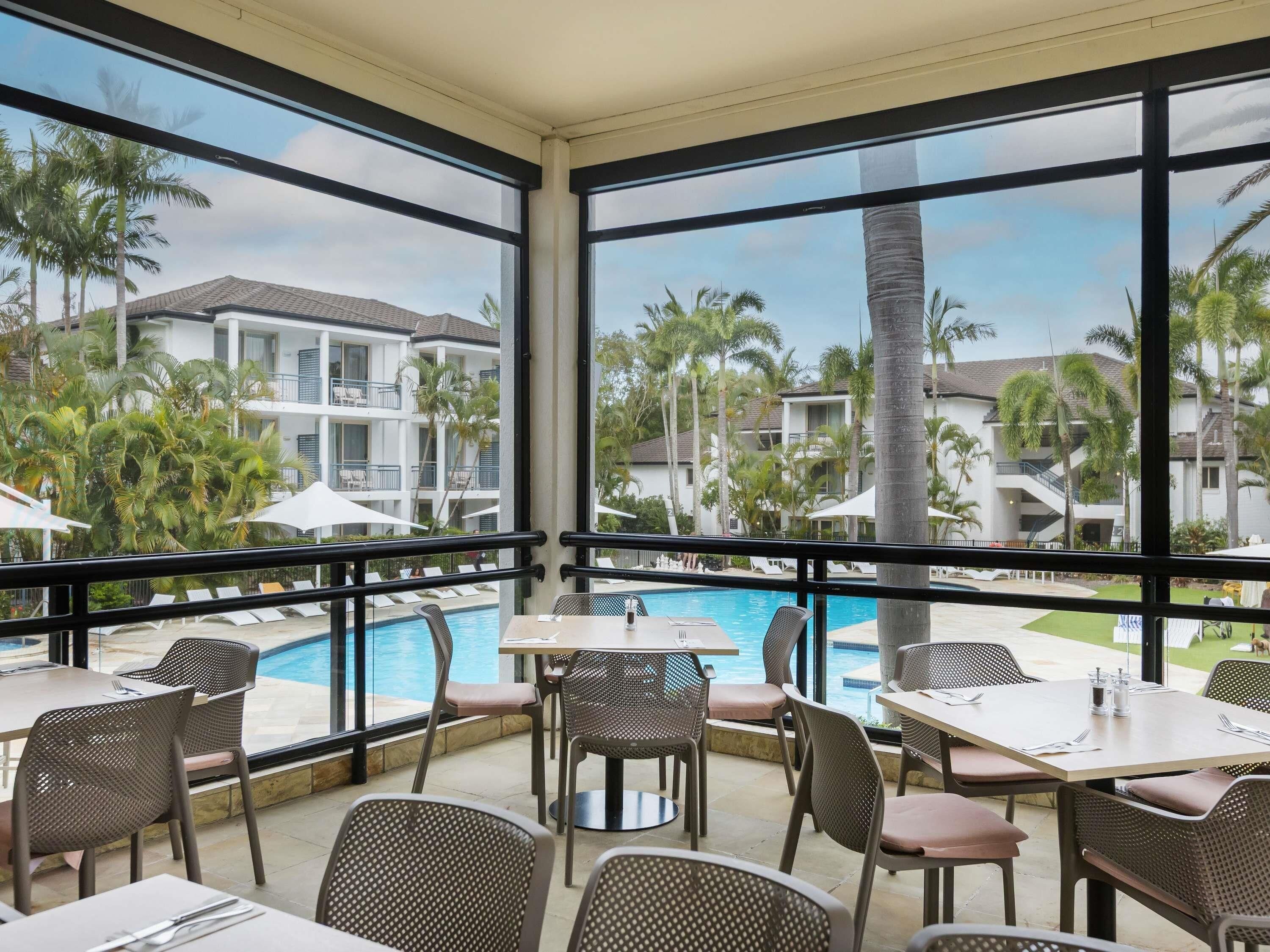 A poolside dining area with tables and chairs, overlooking a swimming pool and a resort with palm trees and buildings.
