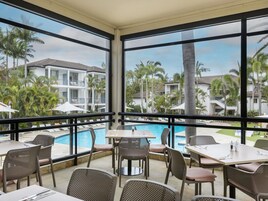 A poolside dining area with tables and chairs, overlooking a swimming pool and a resort with palm trees and buildings.