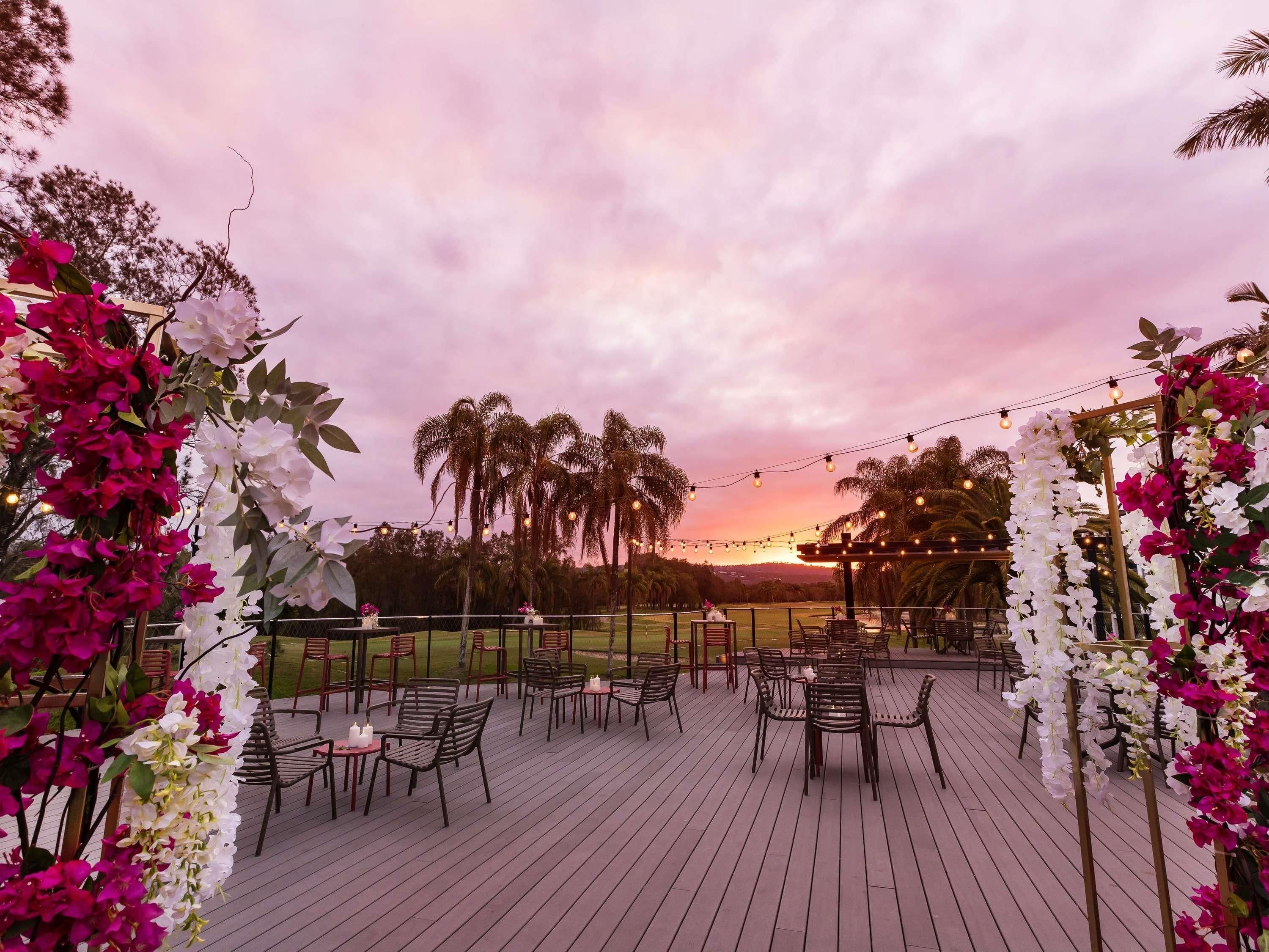 An outdoor patio with tables and chairs, surrounded by flowering plants and palm trees, under a sunset sky.