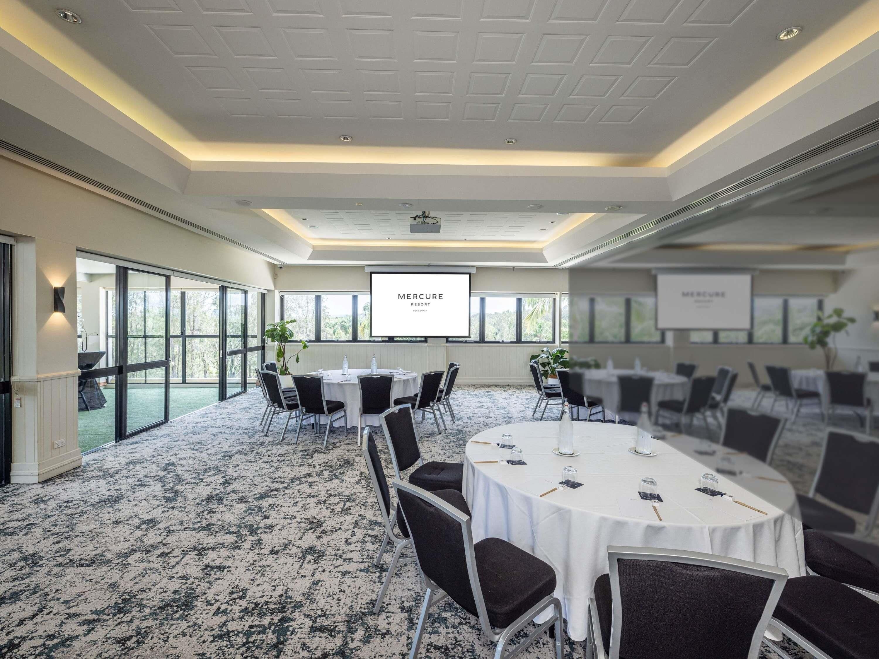 Un salón de conferencias con una pantalla grande, mesas redondas, sillas, piso alfombrado y ventanas amplias.