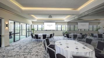 Un salón de conferencias con una pantalla grande, mesas redondas, sillas, piso alfombrado y ventanas amplias.