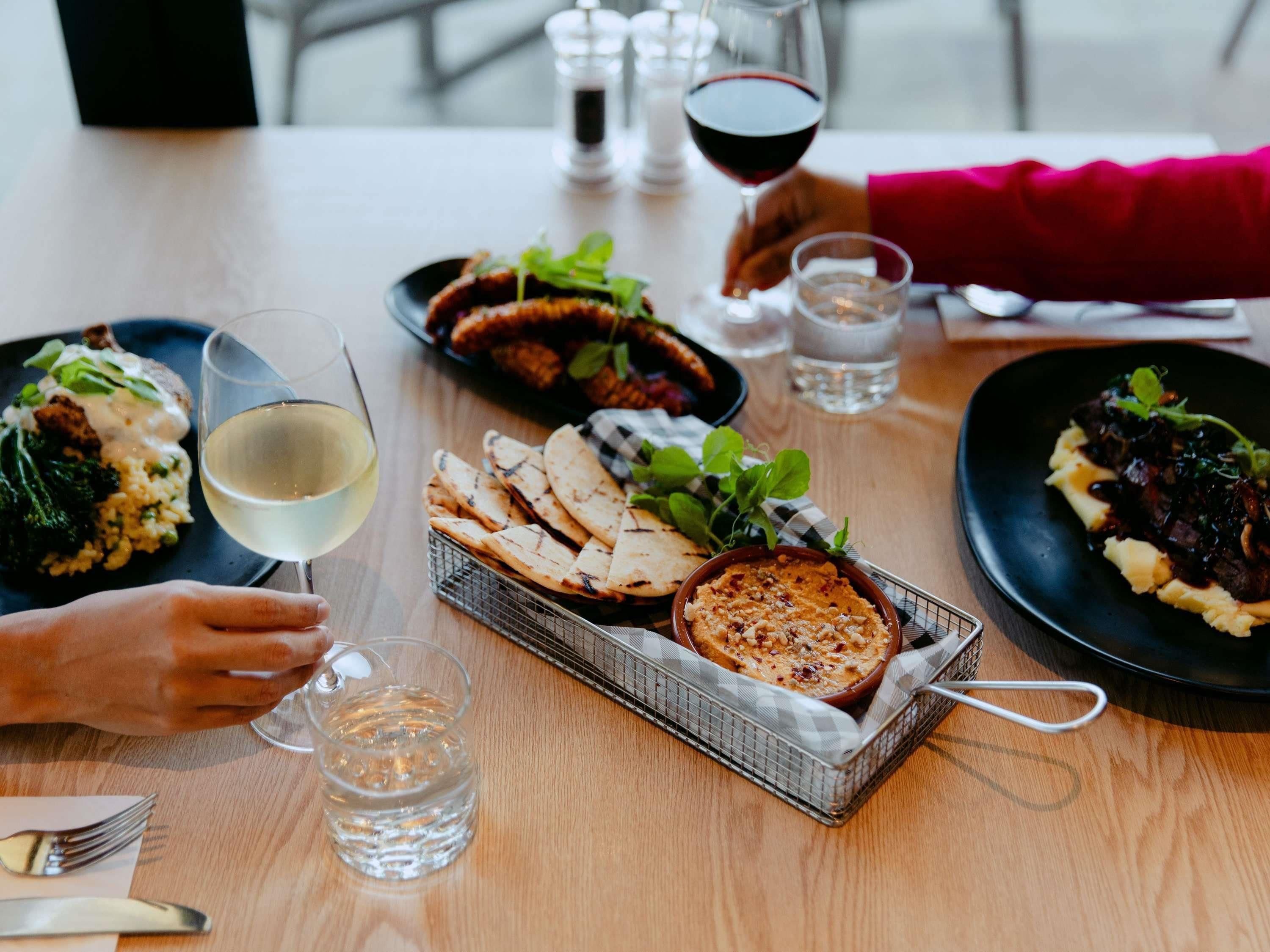 Una mesa de comedor con diversos platos, vasos de agua y vino, y una cesta de pan plano.