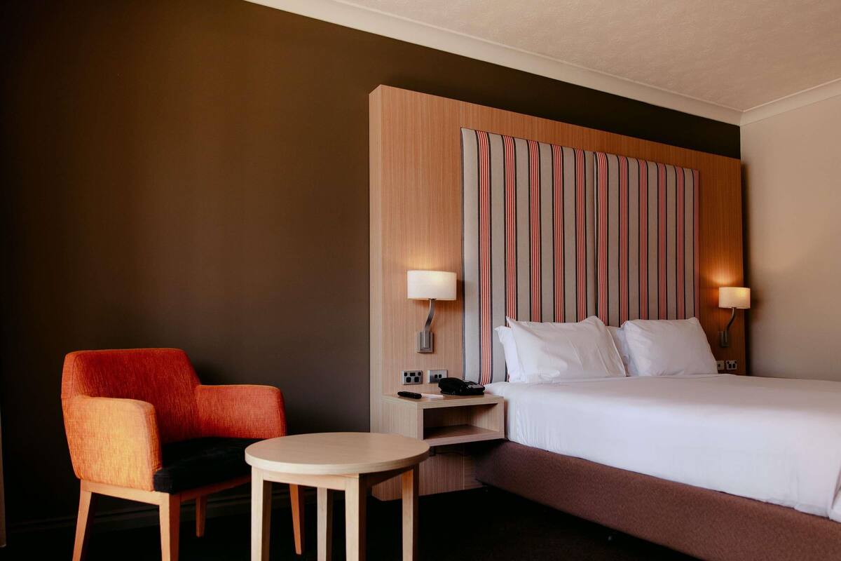 A hotel room with a bed, a chair, a small table, and a nightstand with a lamp.