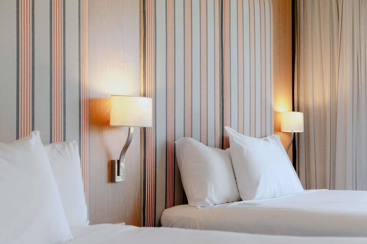 A hotel room with two beds, striped wallpaper, and wall-mounted lamps.
