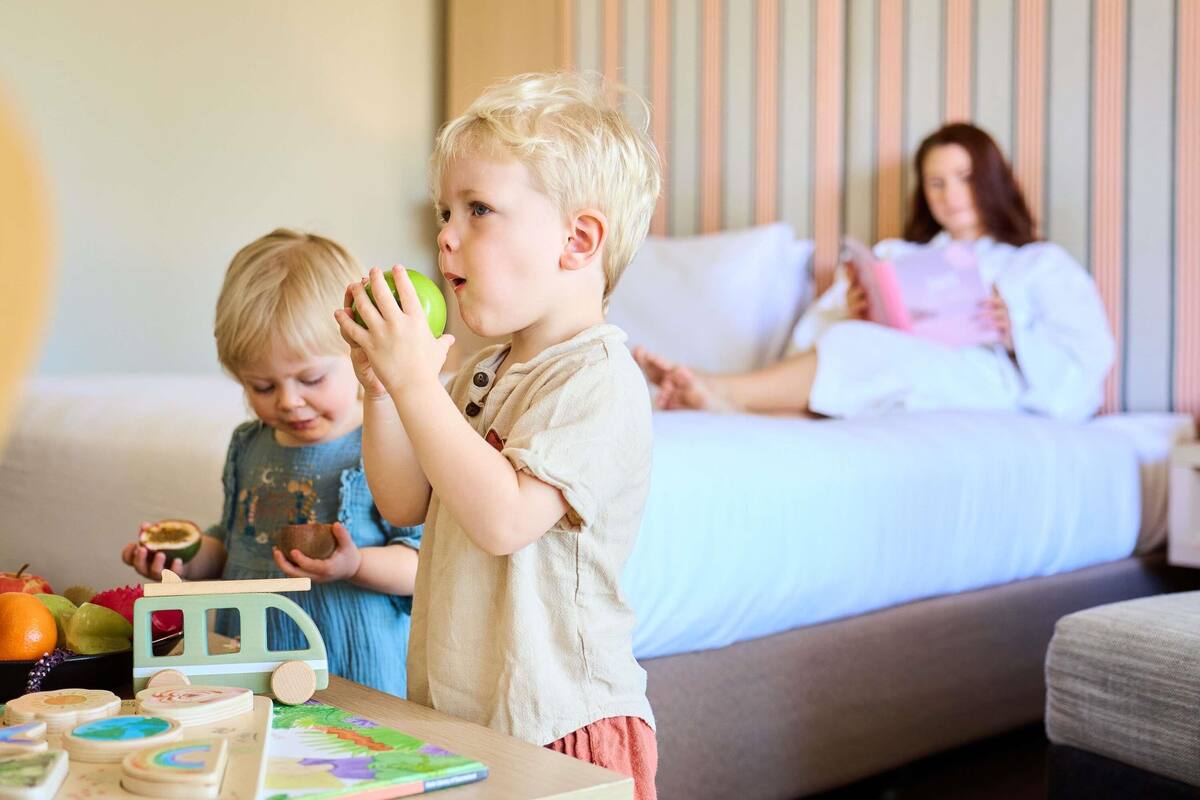 A child holding an apple in a room with a bed and another child in the background.