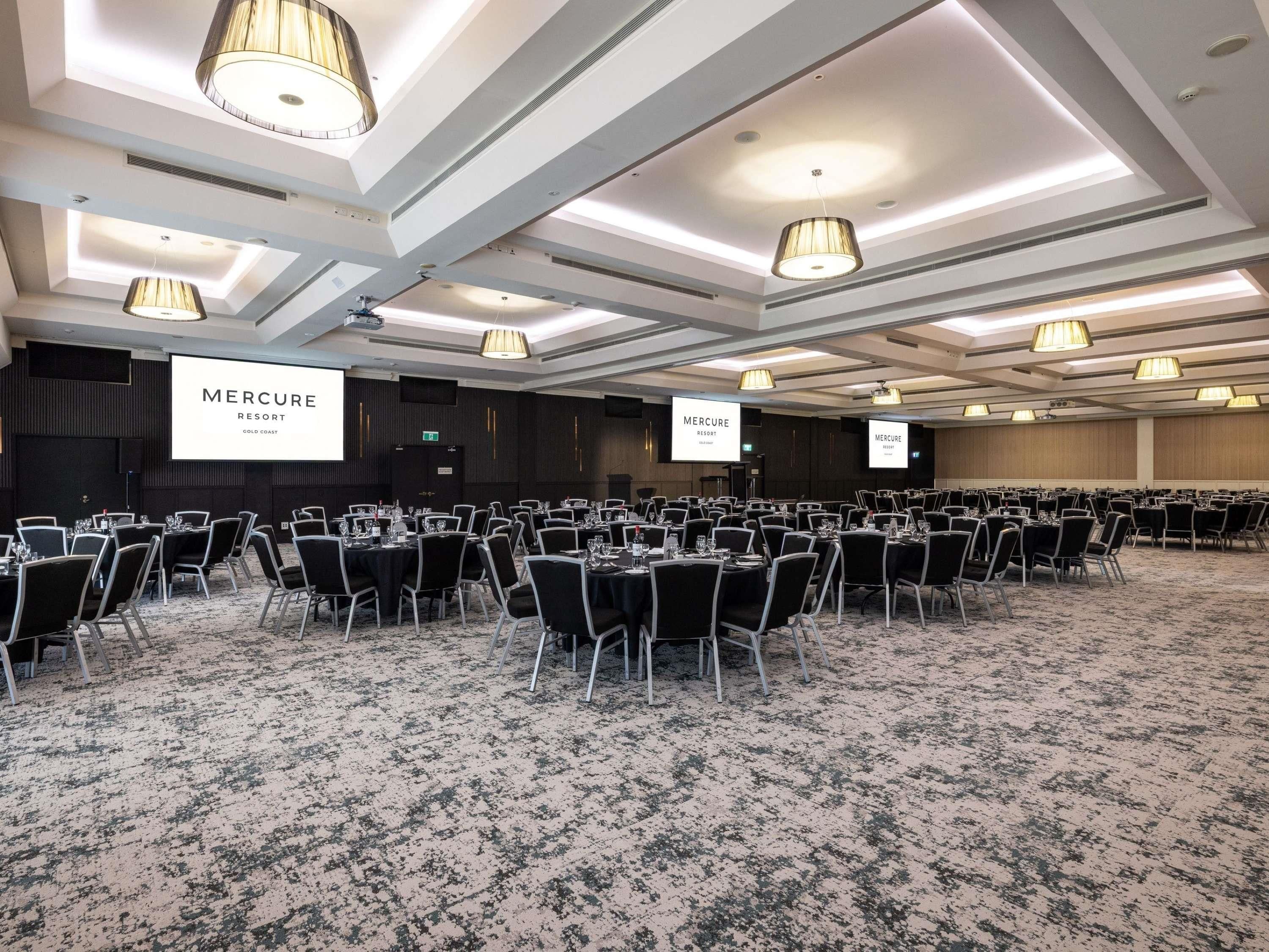 Ein Konferenzraum mit Tischen, Stühlen und großen Bildschirmen, die 'Mercure Resort Gold Coast' zeigen, sowie einem Teppichboden.