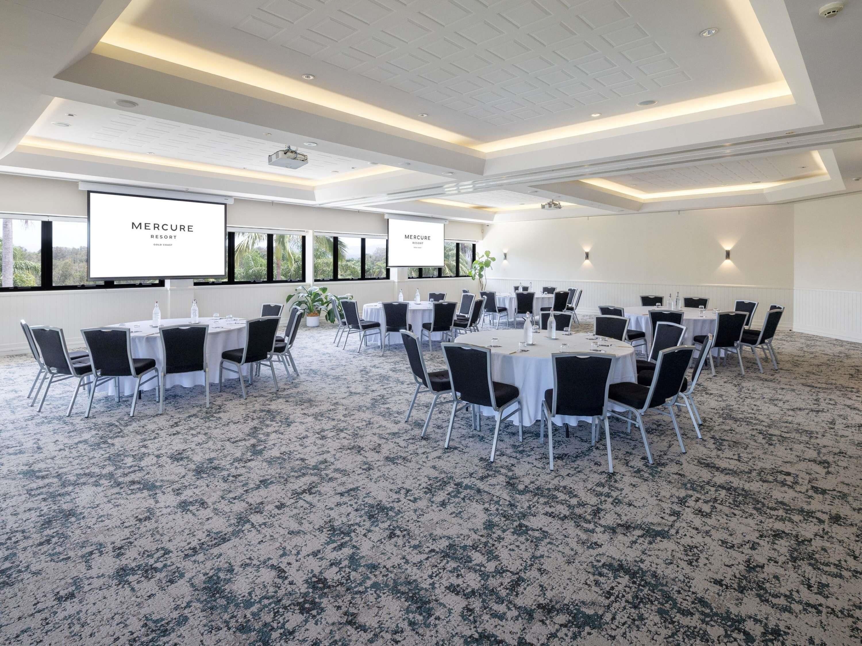 Un salón de conferencias con mesas redondas, sillas, una pantalla de proyector y un amplio ventanal.