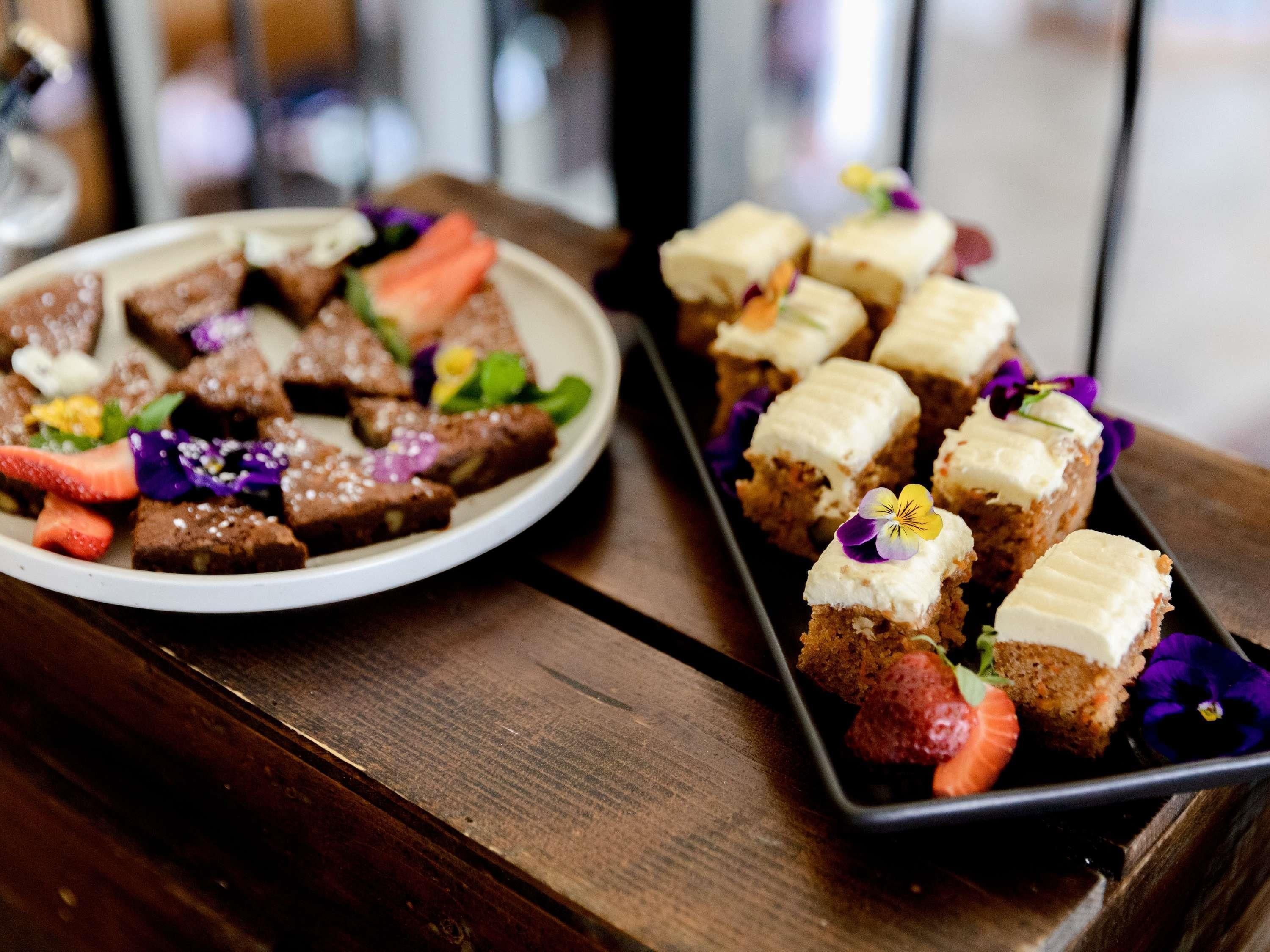 A wooden table with two plates of food, one with brownies and strawberries, the other with cupcakes and flowers.