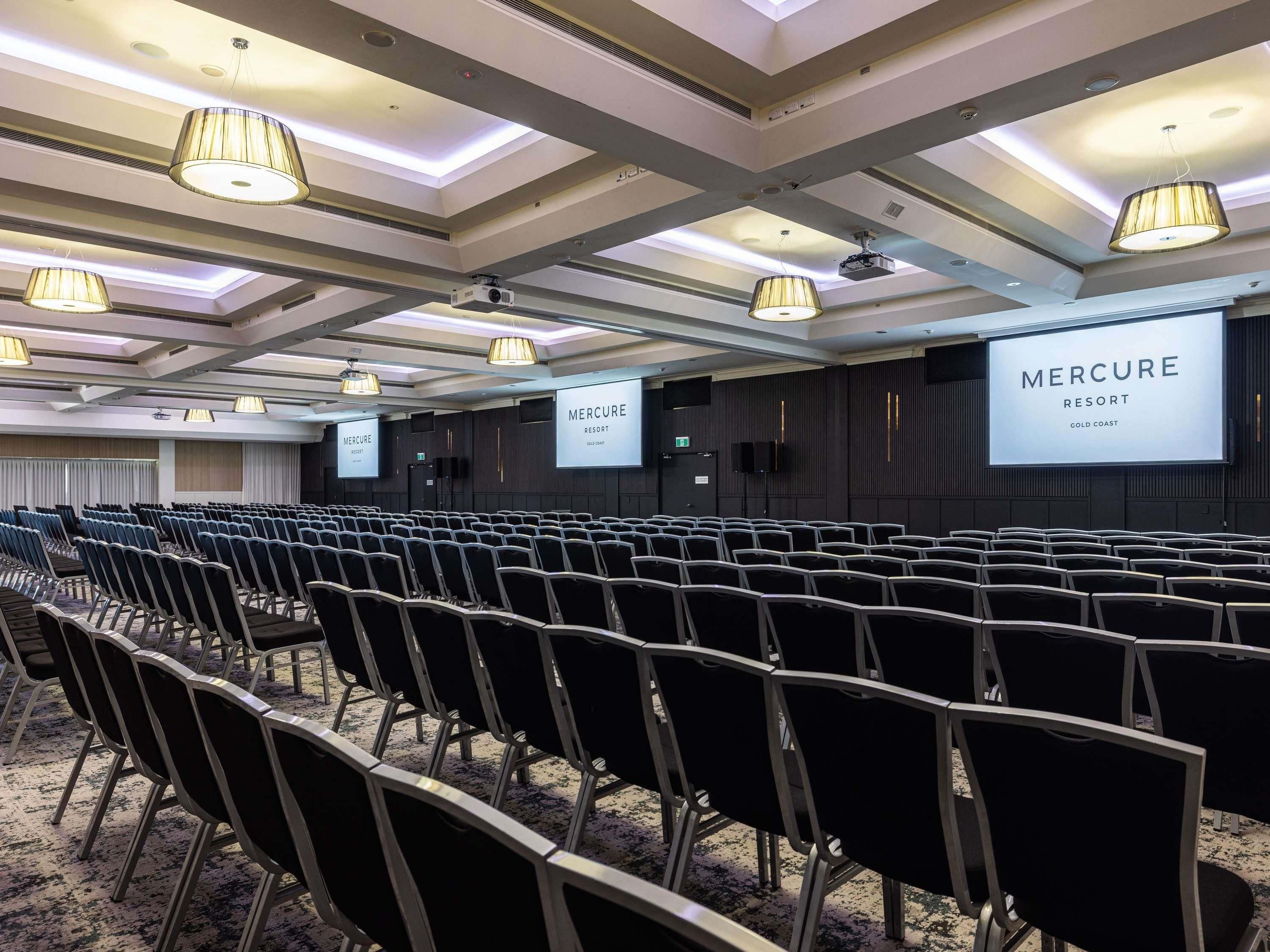 A conference room with rows of chairs, ceiling lights, and a large screen displaying 'MERCURE RESORT GOLD COAST'.
