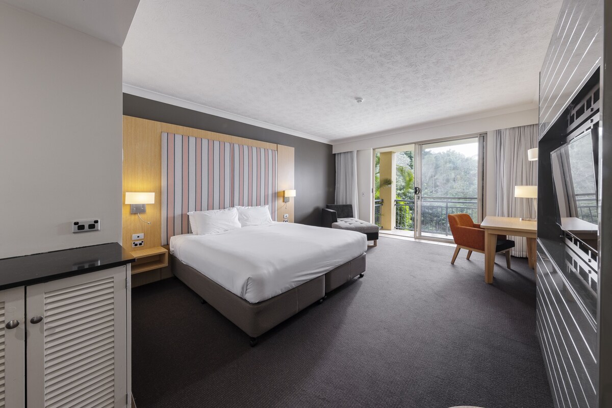 A modern hotel room with a large bed, a desk, and a chair. There is a balcony with a view of greenery.