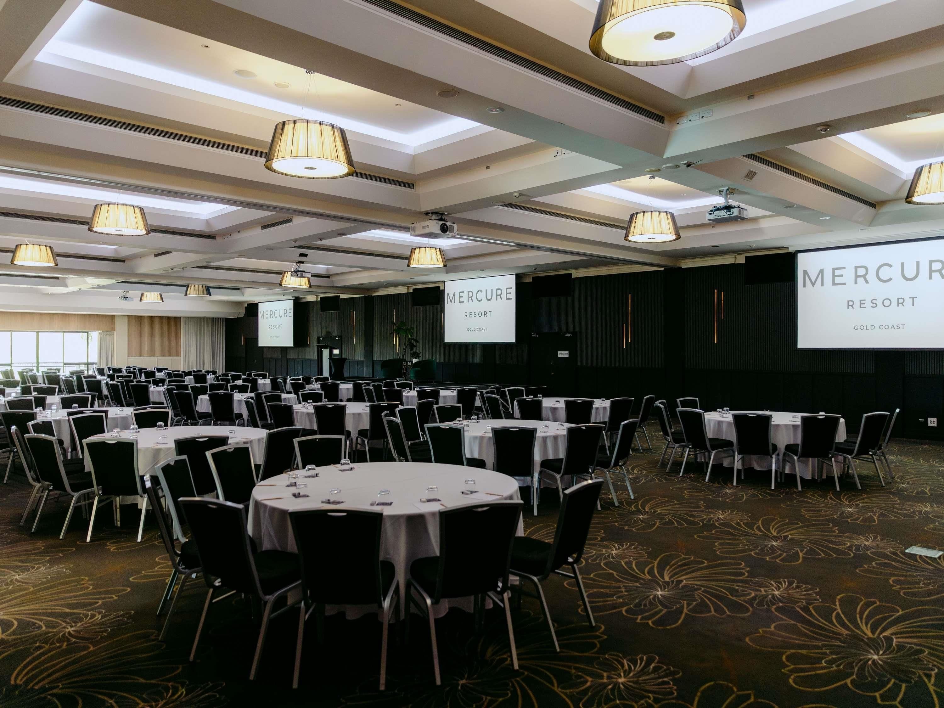 A conference room with round tables, black chairs, and a large screen displaying 'Mercure Resort Gold Coast'.