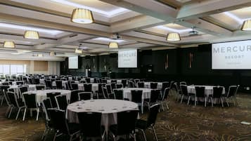 A conference room with round tables, black chairs, and a large screen displaying 'Mercure Resort Gold Coast'.