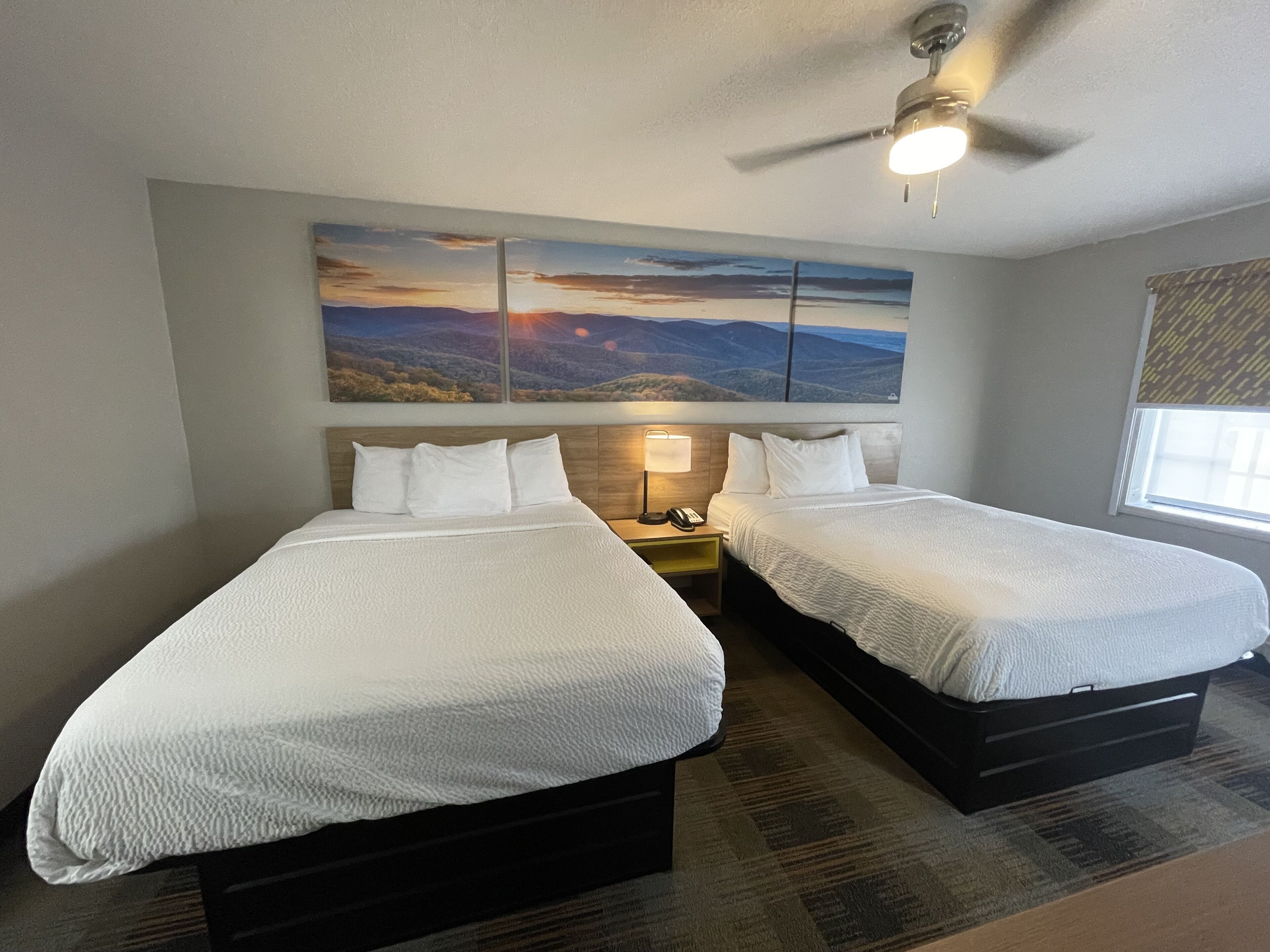 A hotel room with two beds, a ceiling fan, a nightstand with a lamp, and a large landscape painting on the wall.