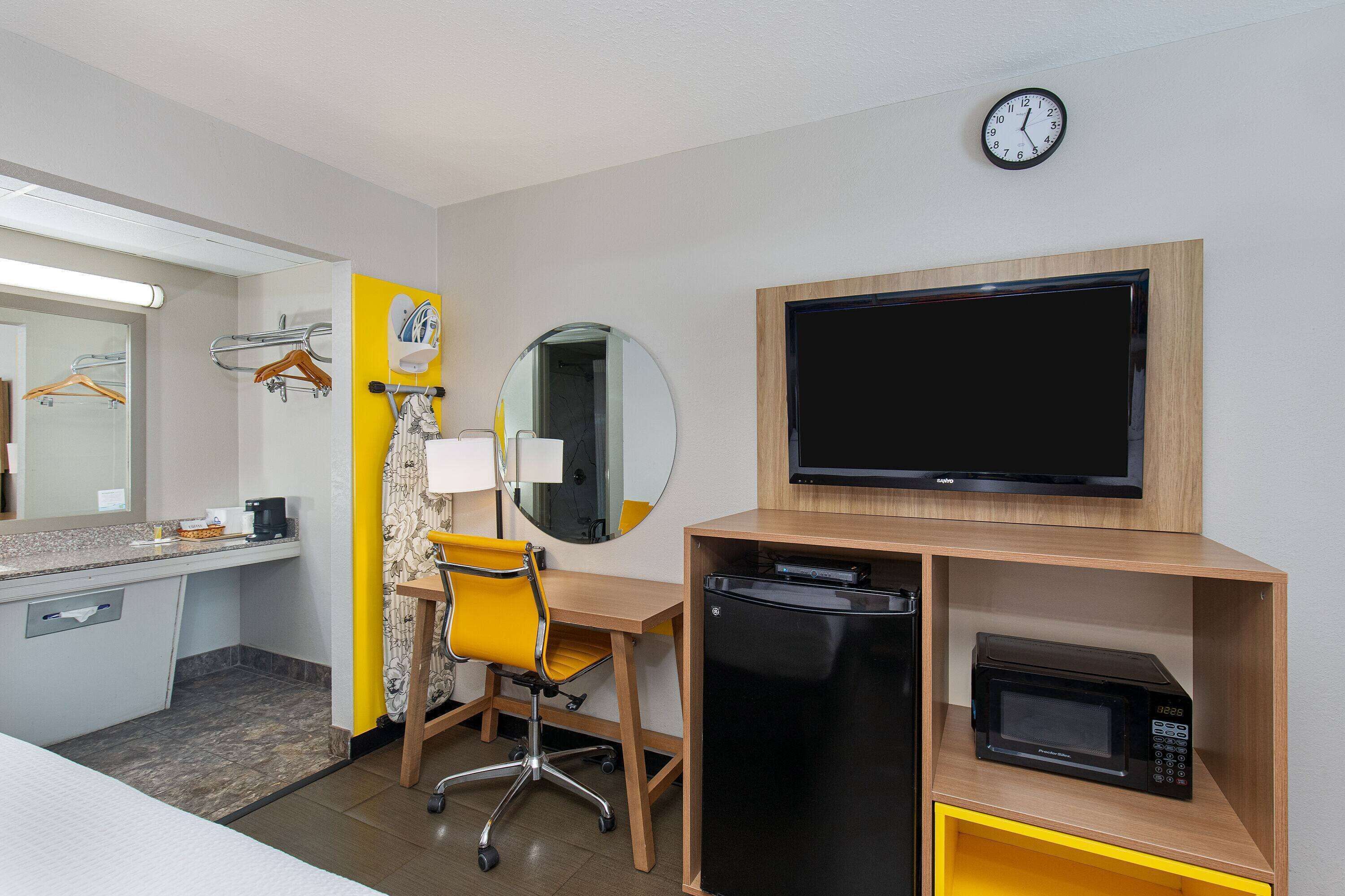 A hotel room with a desk, chair, microwave, TV, and a clock on the wall.