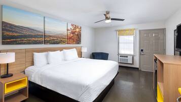 A hotel room with a large bed, a desk, a chair, a ceiling fan, and artwork on the wall.