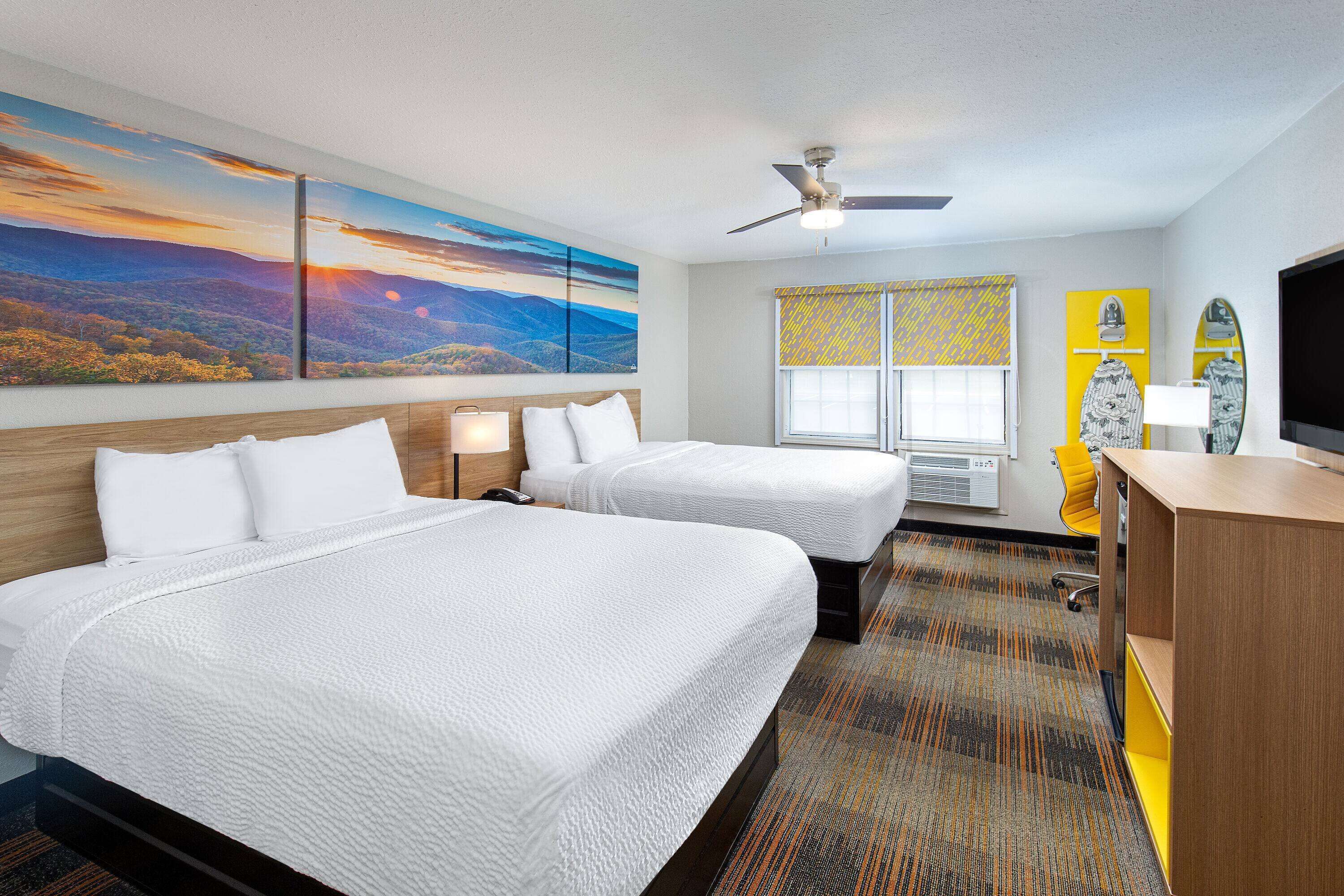A hotel room with two beds, a ceiling fan, a large landscape painting, a window with yellow blinds, and a television on a wooden cabinet.