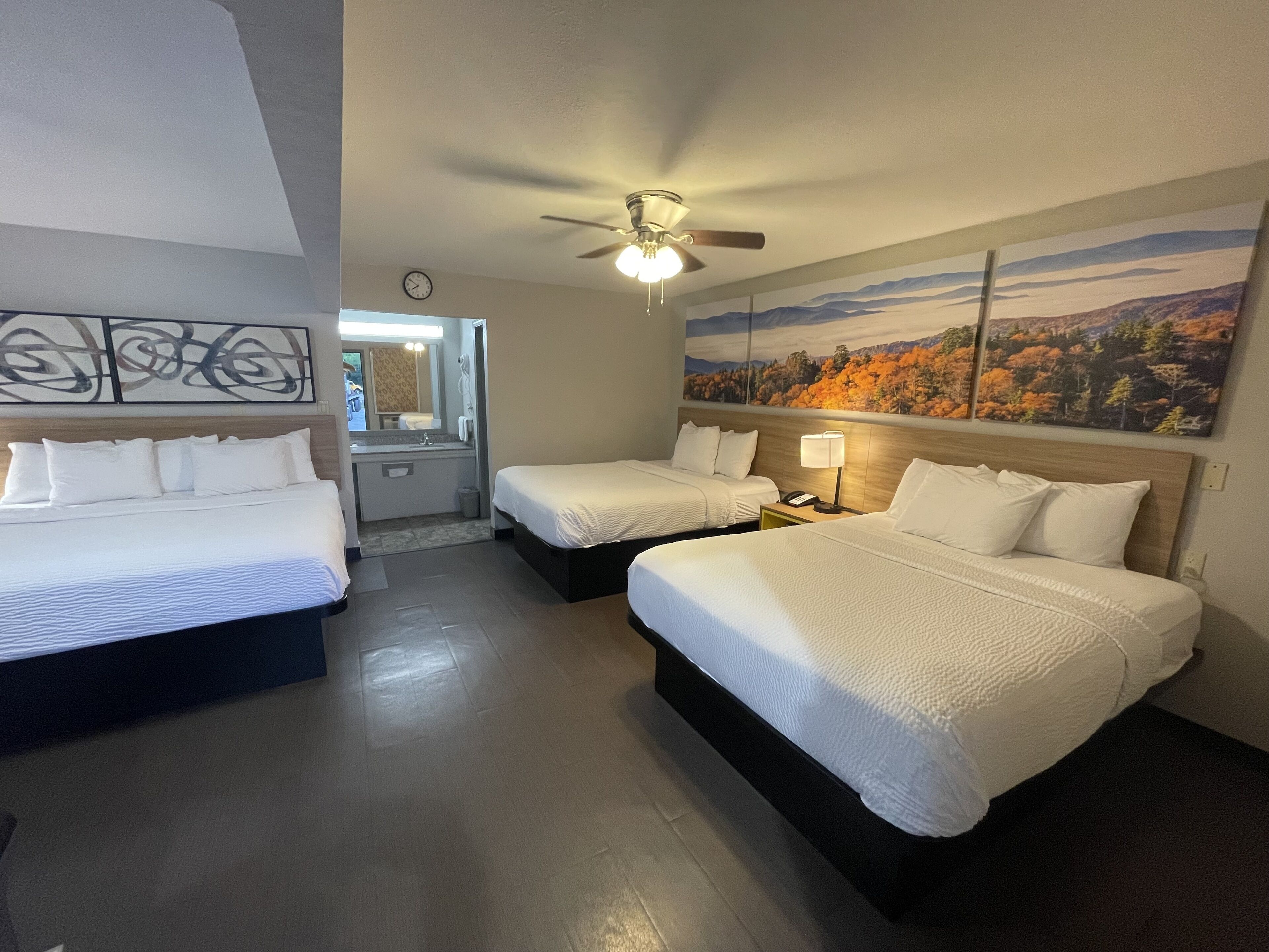 A hotel room with two beds, a ceiling fan, a painting of autumn foliage, and a bathroom visible through an open door.