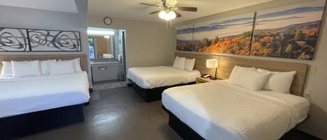 A hotel room with two beds, a ceiling fan, a painting of autumn foliage, and a bathroom visible through an open door.