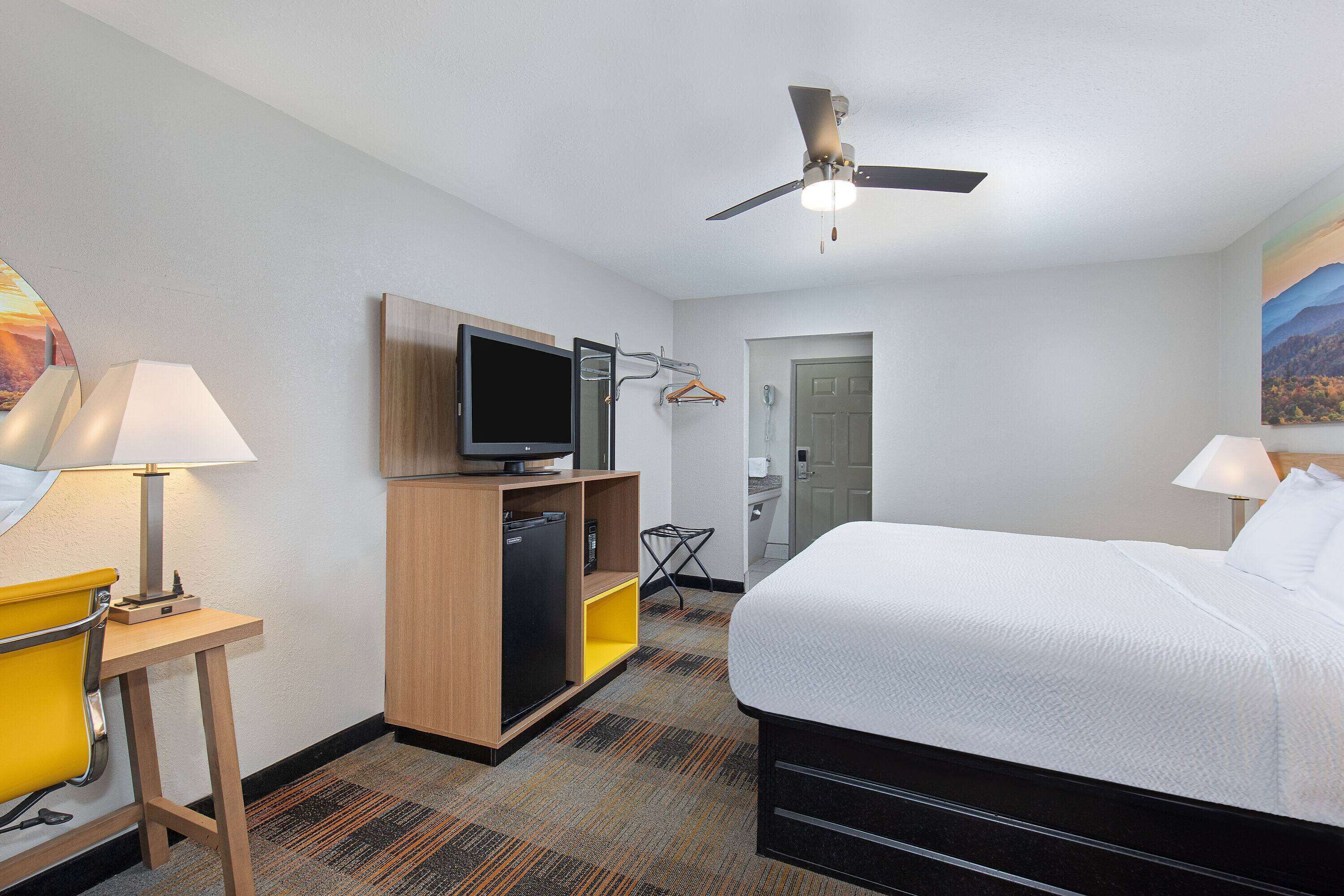 A hotel room with a bed, a television, a desk with a lamp, a wardrobe, and a ceiling fan.