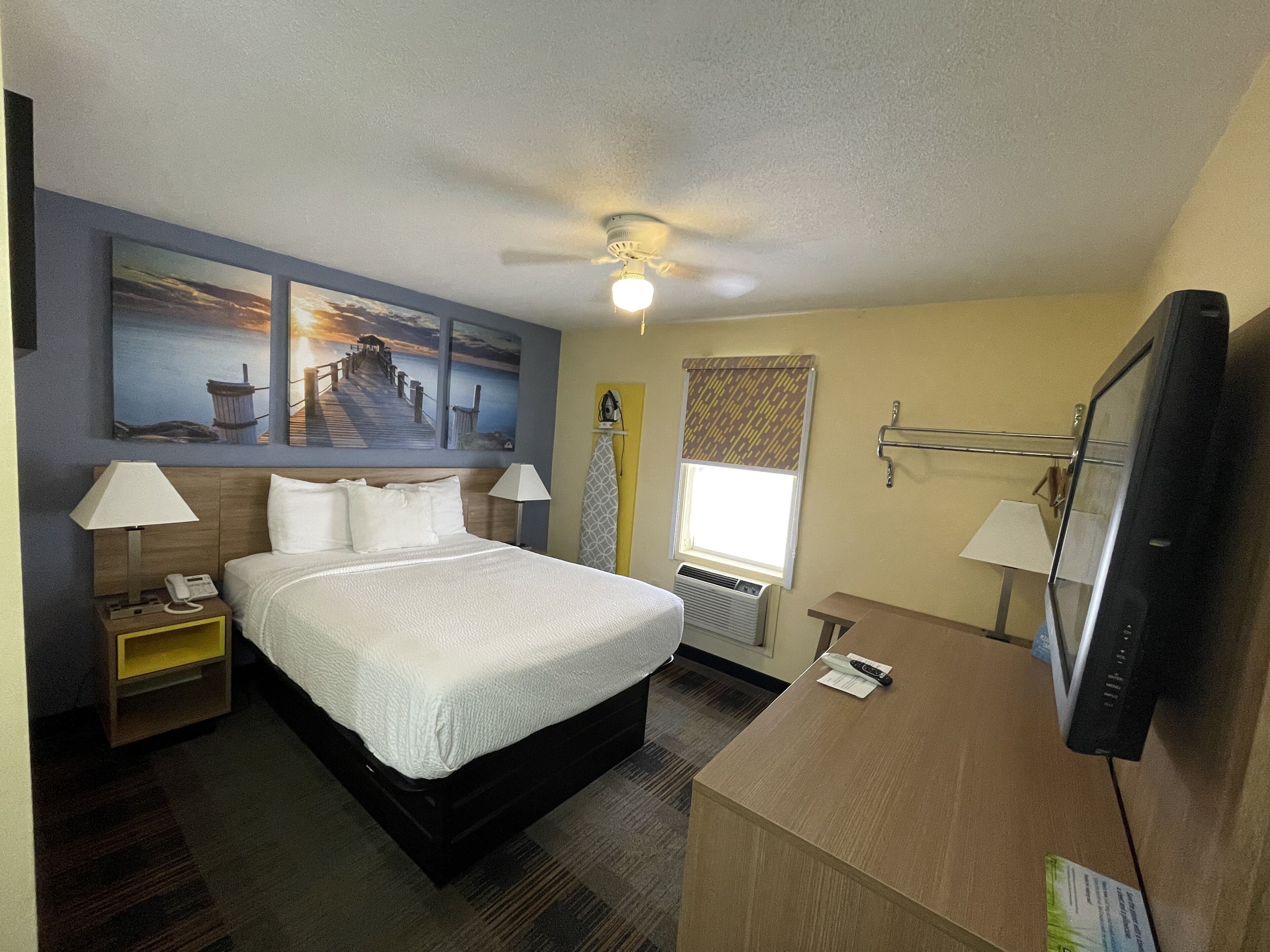 A hotel room with a bed, bedside tables, a desk with a computer, a ceiling fan, and artwork on the walls.