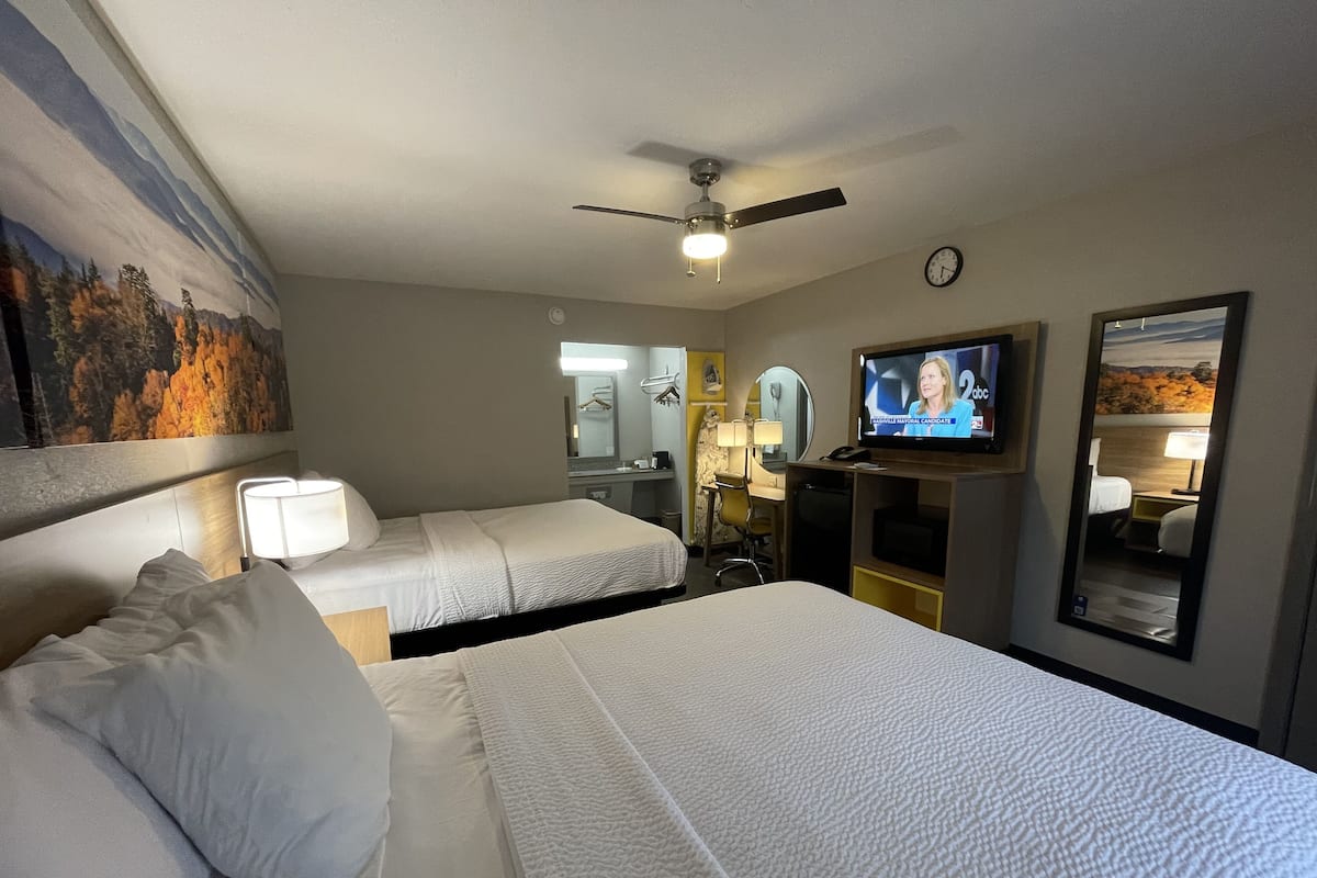 A hotel room with two beds, a ceiling fan, a TV, a clock, and a painting on the wall.